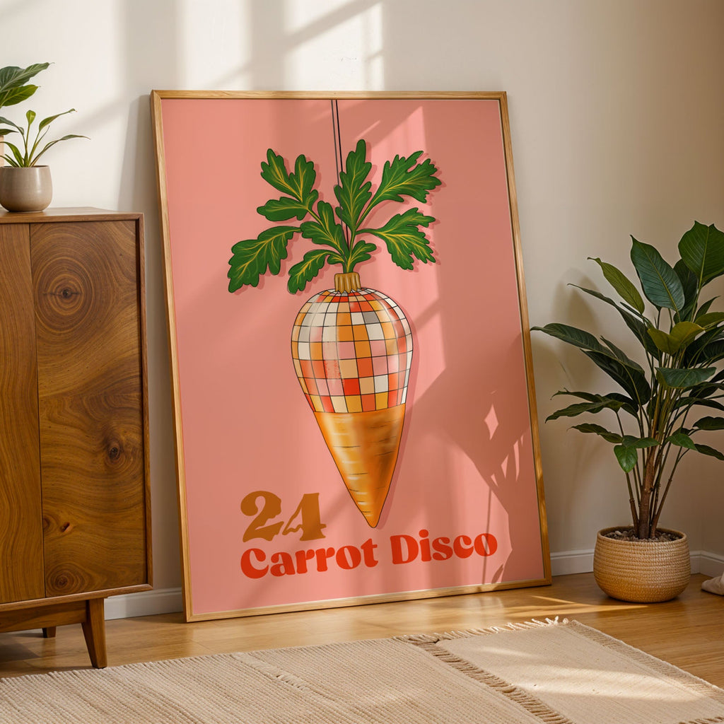 A framed retro-style wall art print leaning against a wall in a styled room. The print shows a carrot-shaped disco ball with green leaves on a pink background and the text '24 Carrot Disco.