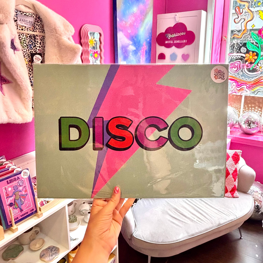 A person holds a square wall art print featuring the word 'DISCO' in large multicolored letters over a pink and purple lightning bolt on a muted sage background. The photo is taken in a bright pink room with shelves, decor, and a white lounge chair visible.