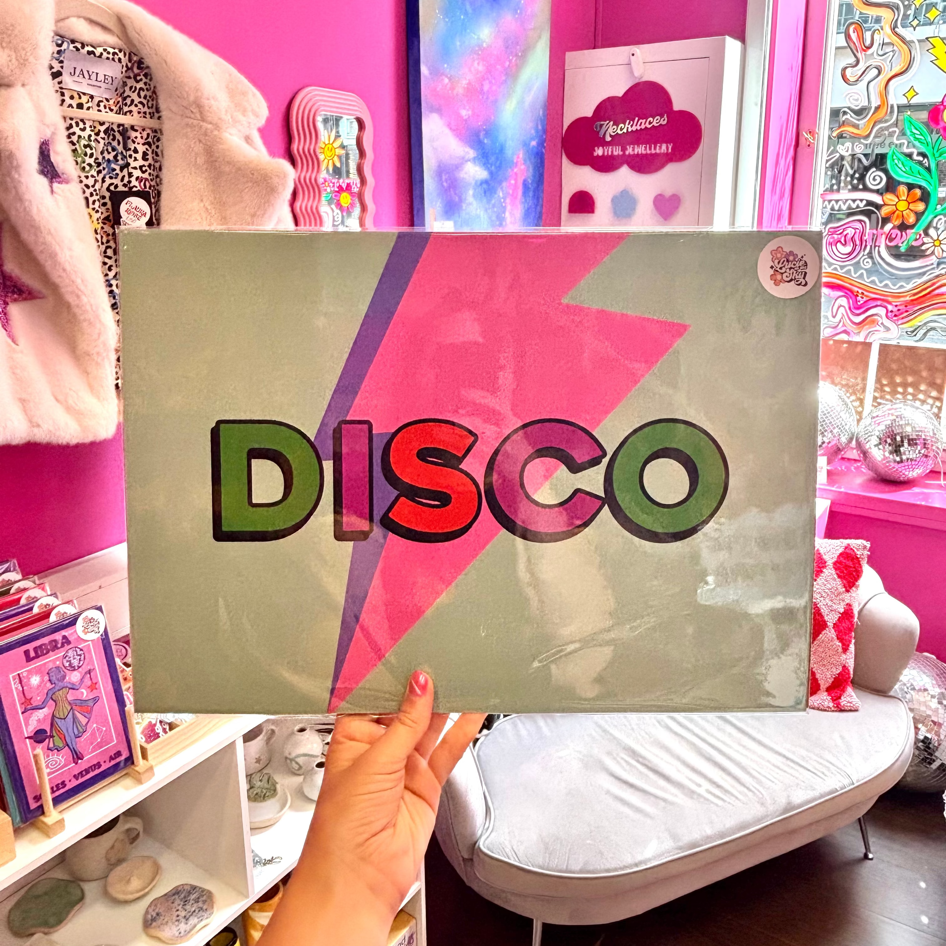 A person holds a square wall art print featuring the word 'DISCO' in large multicolored letters over a pink and purple lightning bolt on a muted sage background. The photo is taken in a bright pink room with shelves, decor, and a white lounge chair visible.
