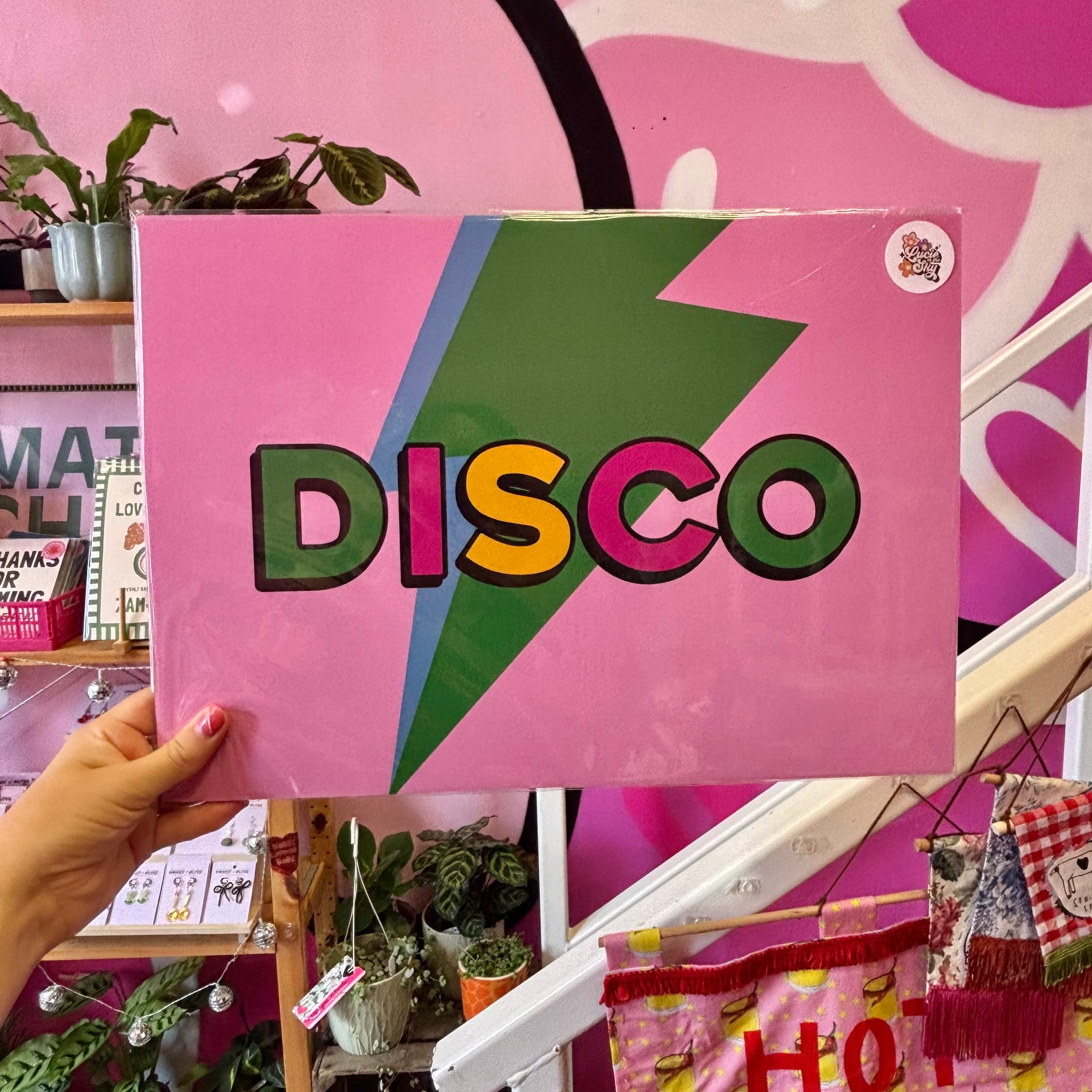 A hand holds a square pink sign featuring a large green lightning bolt and the word 'DISCO' in colorful letters, set against a pink and black illustrated wall with shelves and plants in the background.