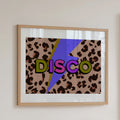 A framed art print featuring a retro 1970s-style 'DISCO' graphic with bold multicolored letters over a leopard print background and a purple lightning bolt, hung on a light beige wall.