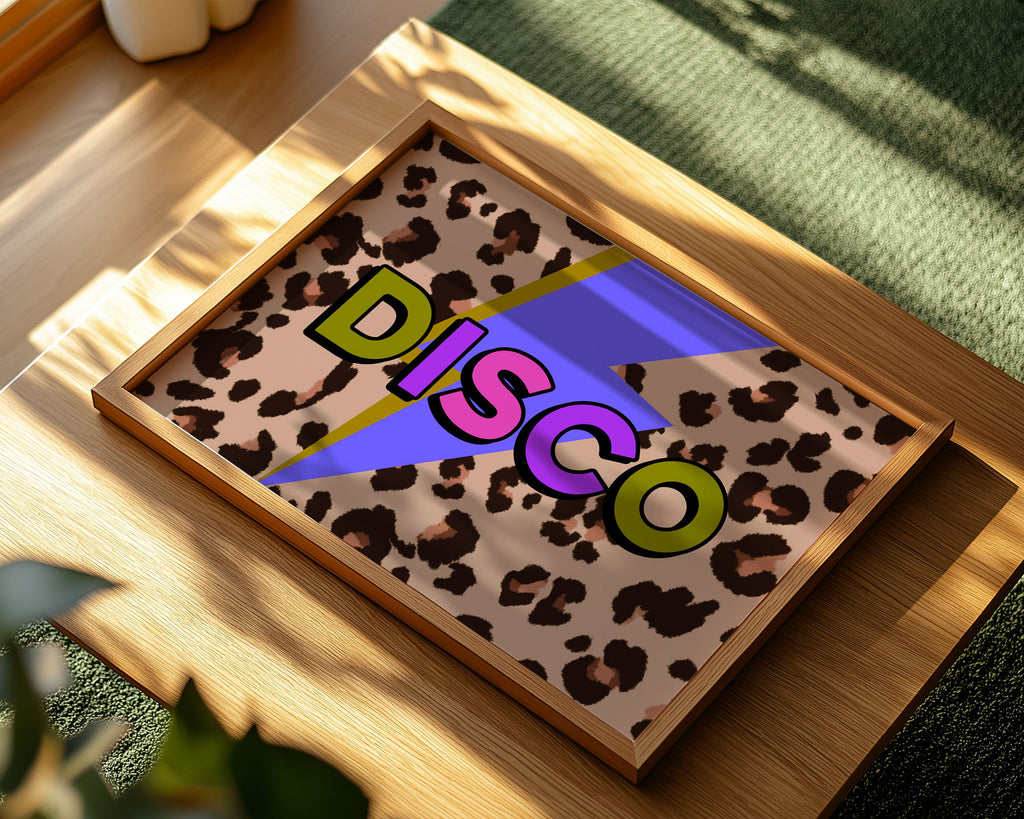A framed square art print featuring a leopard print background with a purple and blue lightning bolt and the word 'DISCO' in bold retro letters, displayed on a wooden surface in warm sunlight.