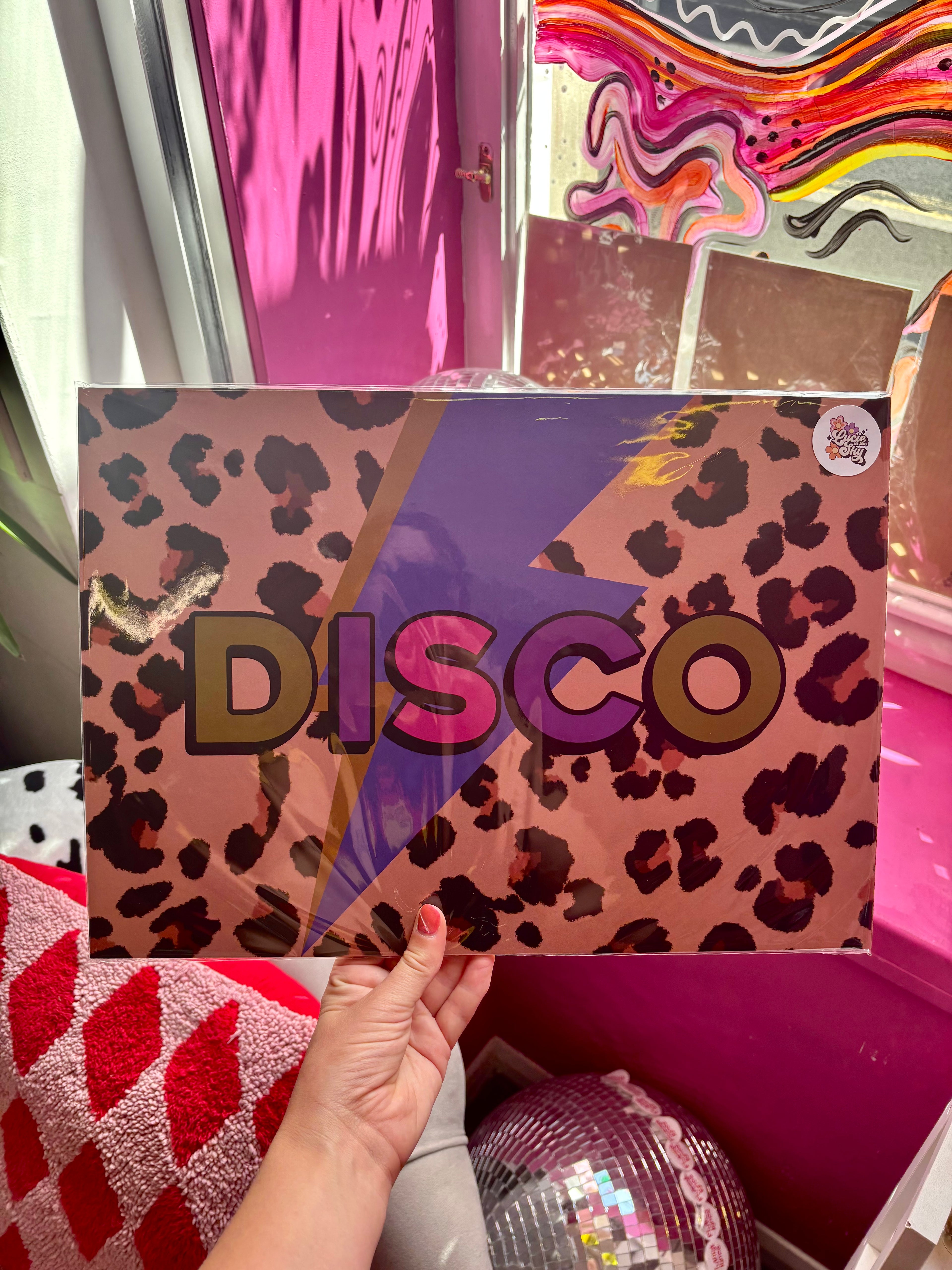 A hand holds a square box or package with a pink leopard print background, a large purple lightning bolt, and the word 'DISCO' in bold multicolor letters. The scene is indoors with bright pink decor and a disco ball visible in the background.