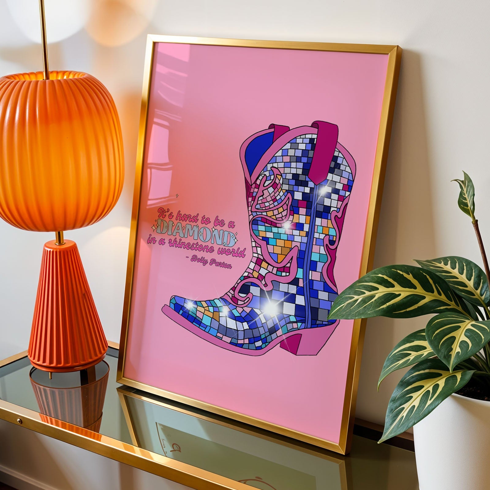 A framed art print featuring a colorful mosaic cowboy boot on a pink background with the quote 'It's hard to be a diamond in a rhinestone world' by Dolly Parton" , styled on a glass-topped side table next to an orange lamp and a green plant.