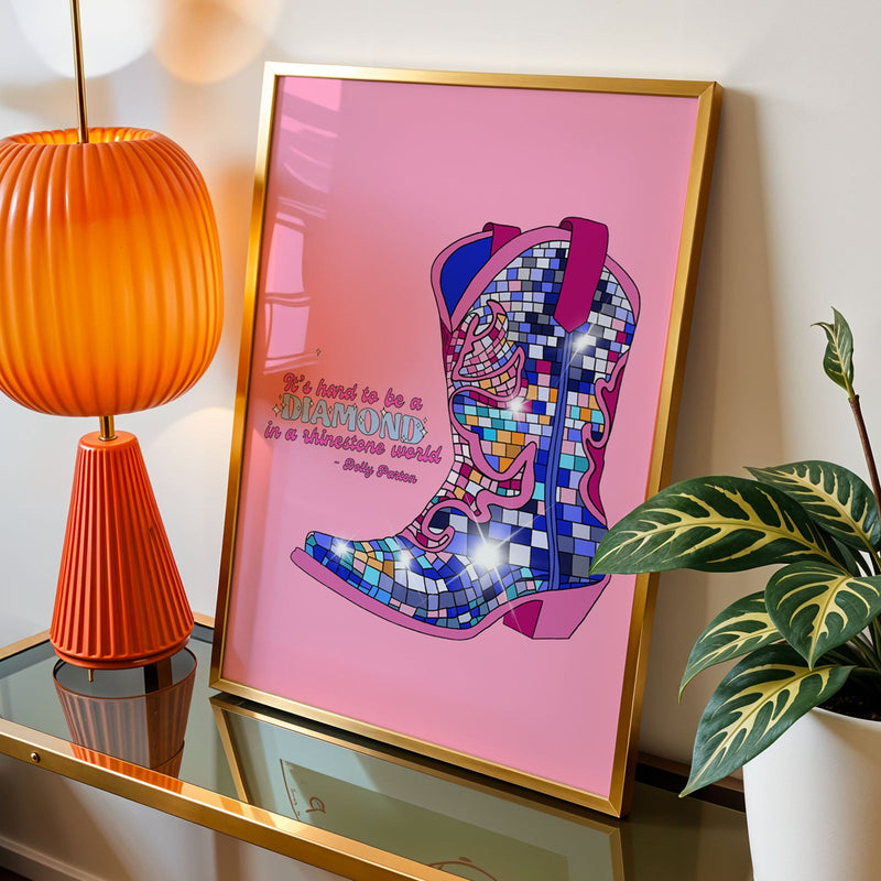 A framed art print featuring a colorful mosaic cowboy boot on a pink background with the quote 'It's hard to be a diamond in a rhinestone world' by Dolly Parton" , styled on a glass-topped side table next to an orange lamp and a green plant.