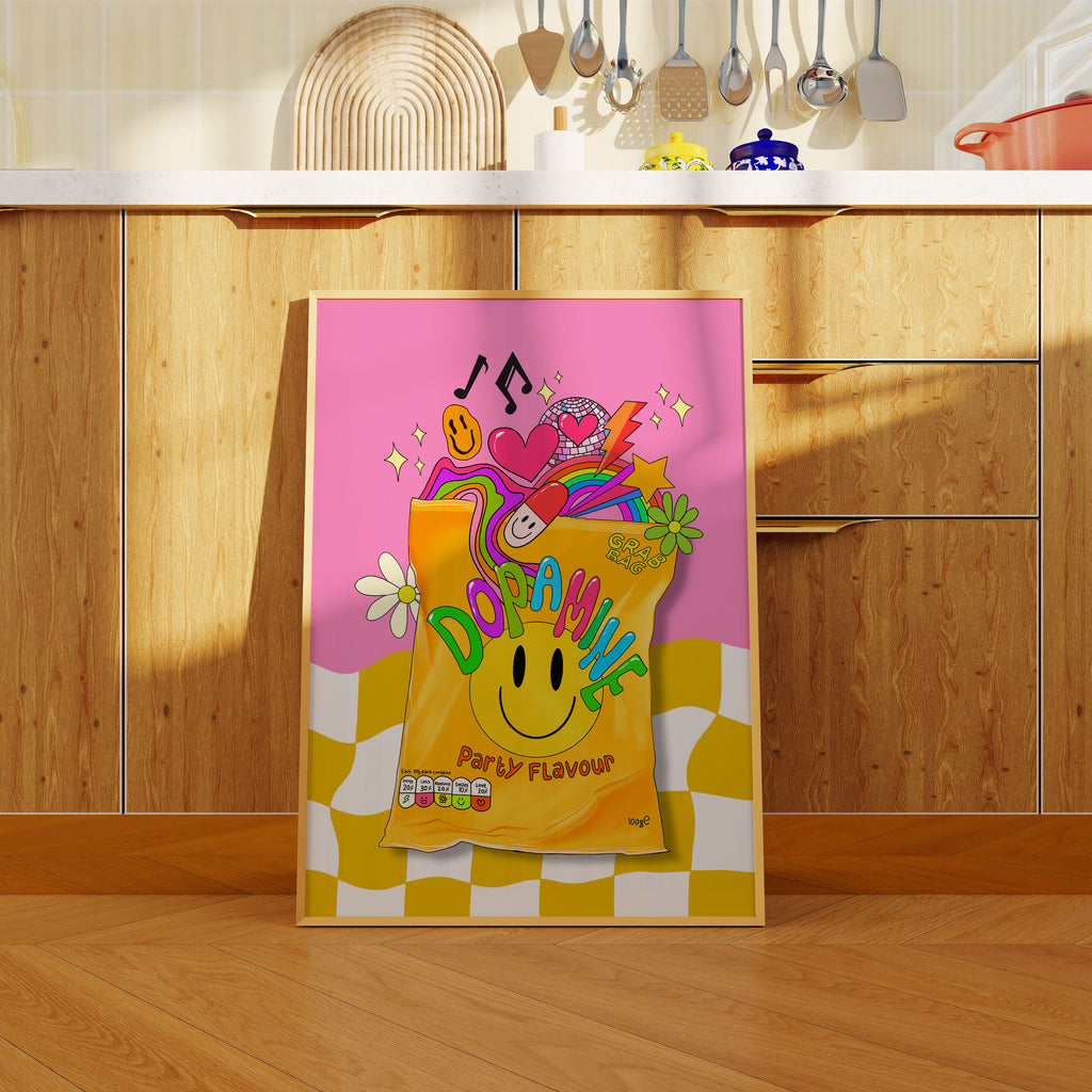 A colorful framed art print leaning against a wooden kitchen cabinet. The print features a pink background with a yellow and white checkered bottom, a yellow smiley-face bag labeled 'DOPAMINE' with 'PARTY FLAVOUR' text, and playful illustrations including a pink heart, a purple snake, disco ball and musical notes.