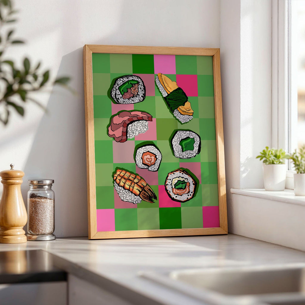 A framed art print featuring colorful illustrated sushi pieces on a green and pink checkered background, leaning against a white kitchen wall on a countertop near a window.