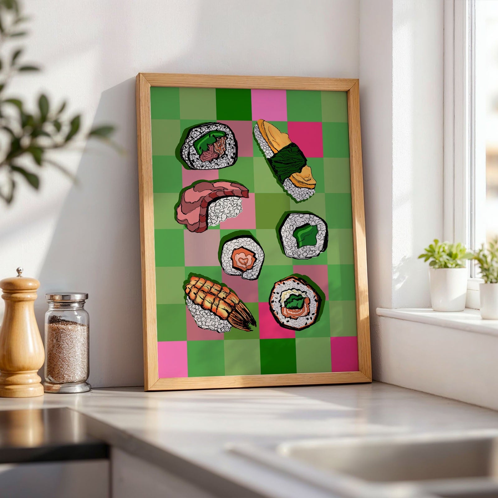 A framed art print featuring colorful illustrated sushi pieces on a green and pink checkered background, leaning against a white kitchen wall on a countertop near a window.