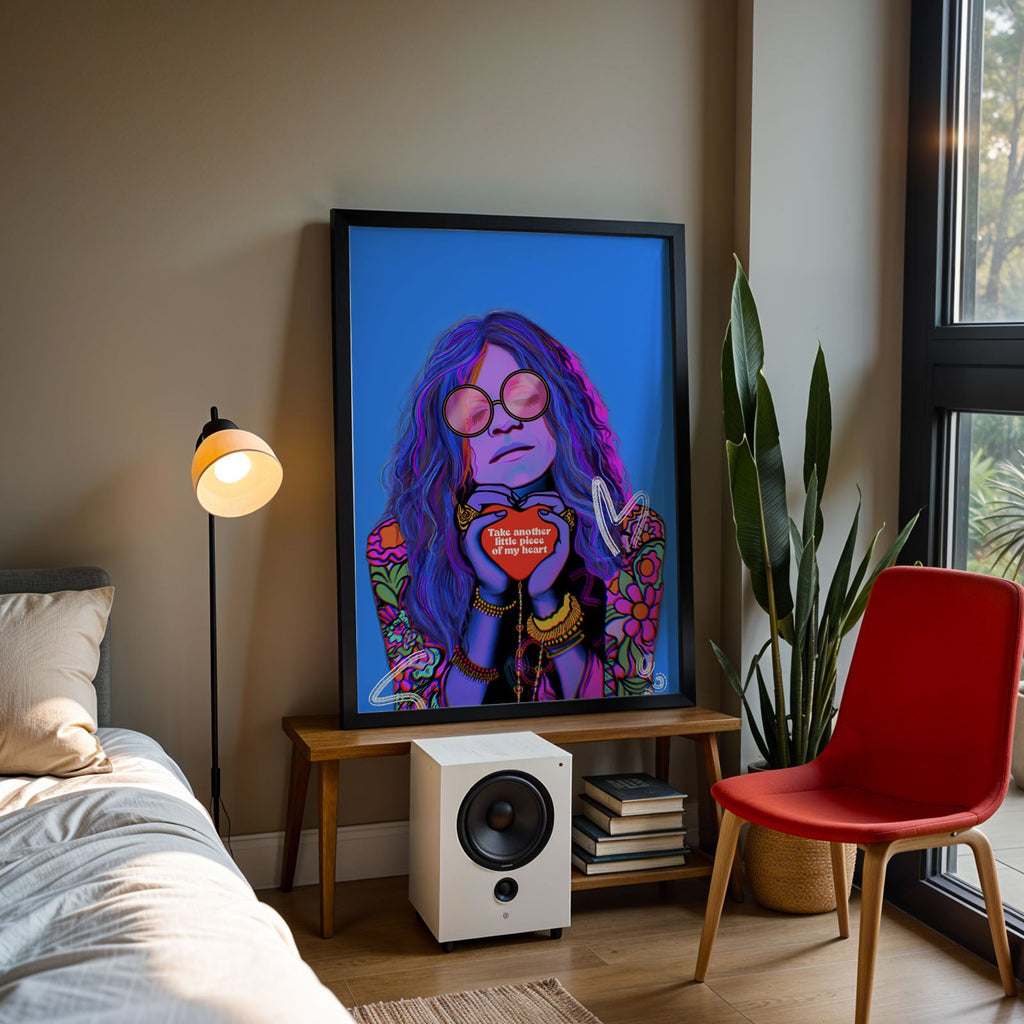A large framed art print featuring a colourful portrait of Janis Joplin with the text 'Take another little piece of my heart' on the heart, displayed in a modern bedroom setting next to a bed, a red chair, and a window.