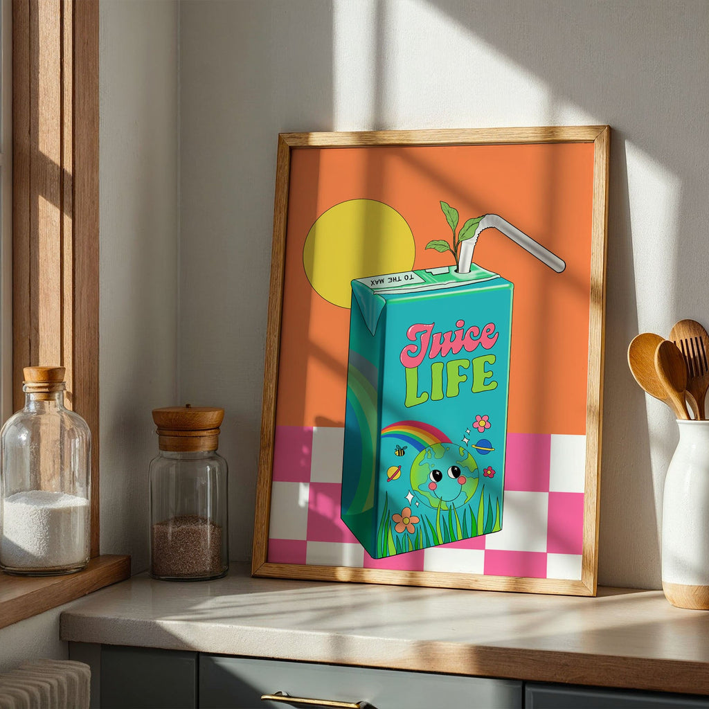 A framed art print leaning on a kitchen counter, featuring a colorful juice carton illustration with the words 'Juice LIFE' against an orange and pink-and-white checkered background. The scene is styled in a sunlit kitchen with jars, wooden utensils, and a plant.