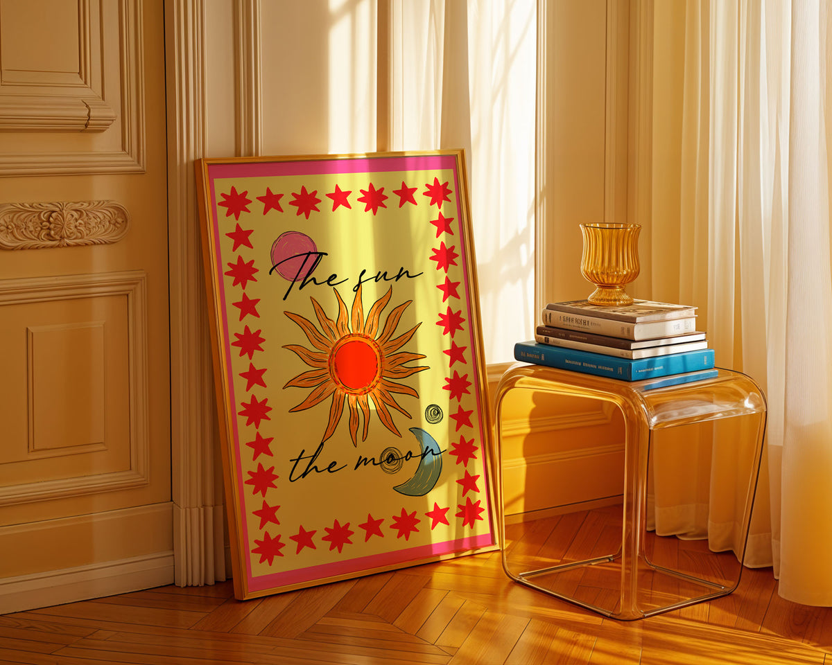 Colourful The Sun & The Moon art print by Lucie In The Sky, featuring an illustrated orange sun, blue crescent moon and red star border on a yellow background, displayed in a gold frame in a sunlit interior.
