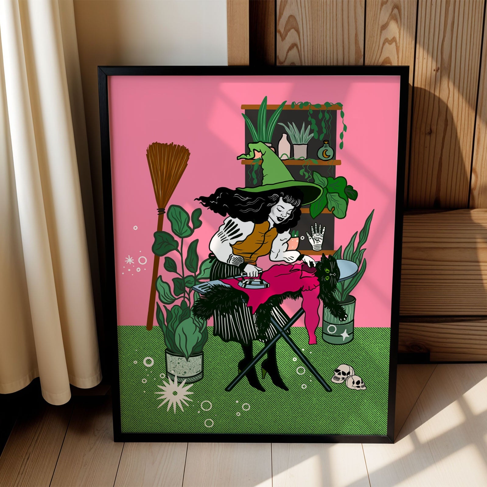 A framed art print leaning against a wooden wall in a sunlit room. The print features a colorful illustration of a witch ironing surrounded by plants, with a bright pink background and green grass at the bottom.