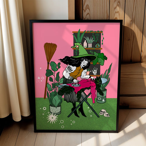 A framed art print leaning against a wooden wall in a sunlit room. The print features a colorful illustration of a witch ironing surrounded by plants, with a bright pink background and green grass at the bottom.