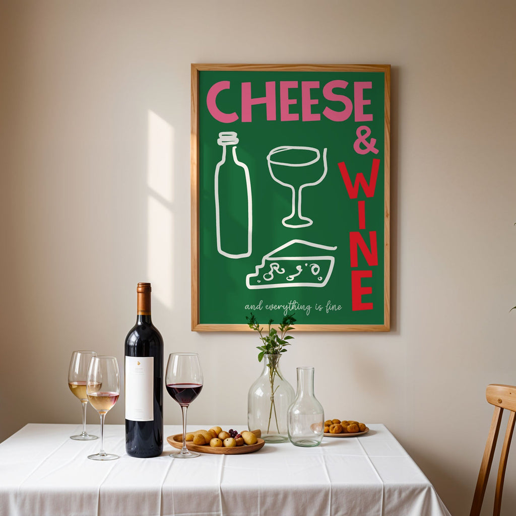 A framed art print featuring a green background with the words 'CHEESE & WINE' in pink and red, along with simple drawings of a bottle, wine glass, cheese wedge, and wine bottle. The print is displayed on a beige wall above a dining table set with a wine bottle, two wine glasses, and small plates of food.