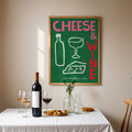 A framed art print featuring a green background with the words 'CHEESE & WINE' in pink and red, along with simple drawings of a bottle, wine glass, cheese wedge, and wine bottle. The print is displayed on a beige wall above a dining table set with a wine bottle, two wine glasses, and small plates of food.